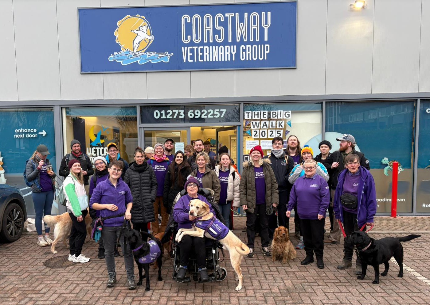 A group of people, some with guide dogs and wearing purple clothing, pose and smile outside the Coastway Veterinary Group building. A sign reads “THE BIG WALK 2025” above the group.