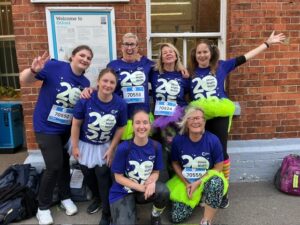 Six women in matching blue “25th Shine Night Walk” shirts and colorful tutus pose and smile together outside, some kneeling and others standing, in front of a brick wall and a sign.