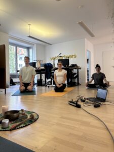 Three people sit on yoga mats in a modern office, meditating. A laptop and ceremonial items are on the floor, and desks with computers are in the background. The word vdpartners is visible on the back wall.