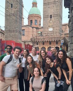 A group of eleven smiling people pose together on a street with historic brick buildings and towers in the background, likely in an Italian city. The atmosphere is cheerful and the sky is overcast.